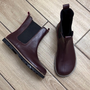 May include: A pair of burgundy Chelsea boots. The boots have a rounded toe, elastic side panels, and a pull tab at the back. The soles are black with a light-colored trim. The boots are made of a smooth leather-like material.
