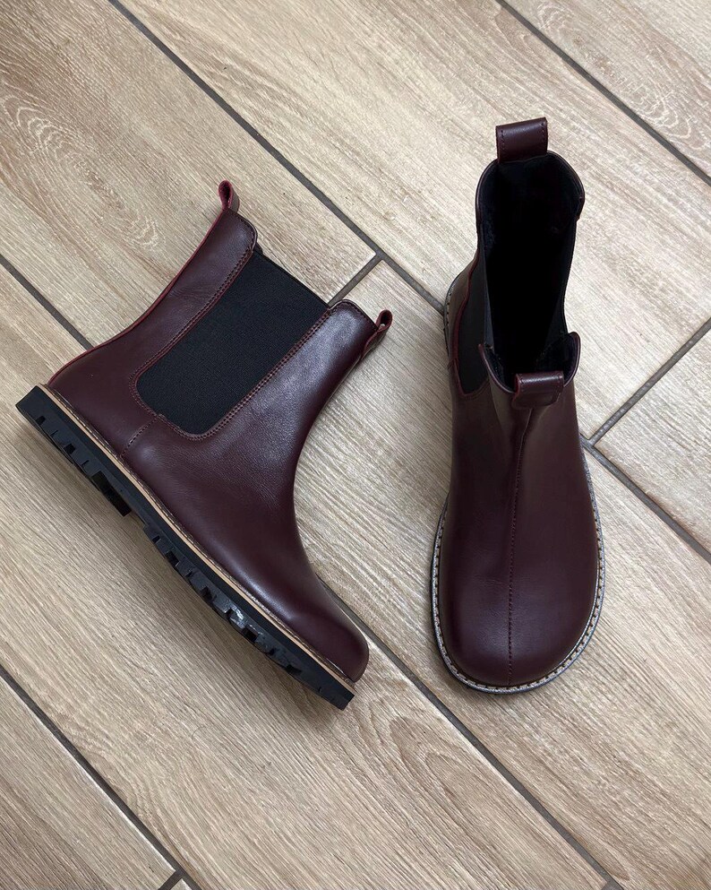 May include: A pair of burgundy Chelsea boots. The boots have a rounded toe, elastic side panels, and a pull tab at the back. The soles are black with a light-colored trim. The boots are made of a smooth leather-like material.
