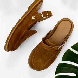 Puede incluir: Dos zuecos de ante marrón con correas ajustables y cierres de hebilla. Los zapatos tienen un diseño de punta redondeada y una suela texturizada. Los zuecos se muestran sobre una superficie blanca con acentos de hojas verdes.