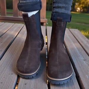 May include: A pair of brown suede Chelsea boots with a rounded toe and a thick, dark gray sole. The boots have a pull tab at the back and are worn with rolled-up jeans. The boots are on a wooden deck.