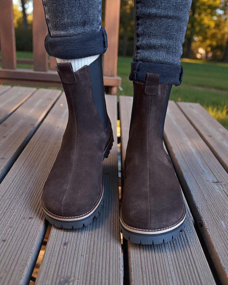 May include: A pair of brown suede Chelsea boots with a rounded toe and a thick, dark gray sole. The boots have a pull tab at the back and are worn with rolled-up jeans. The boots are on a wooden deck.