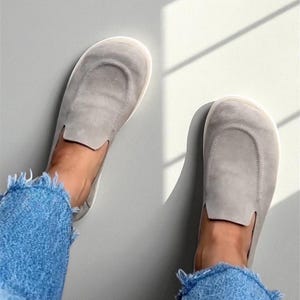 Op de afbeelding: Een paar lichtgrijze instappers met een afgerond teenontwerp. De schoenen zijn gemaakt van een zacht materiaal en worden getoond met een blauwe spijkerbroek met rafelige randen. Bovenaanzicht.