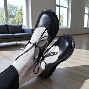 Op de afbeelding: Zwarte leren Mary Jane-schoenen met striksluitingen. De schoenen worden gedragen met witte sokken met een patroon. De foto is binnenshuis genomen met natuurlijk licht en een houten vloer.