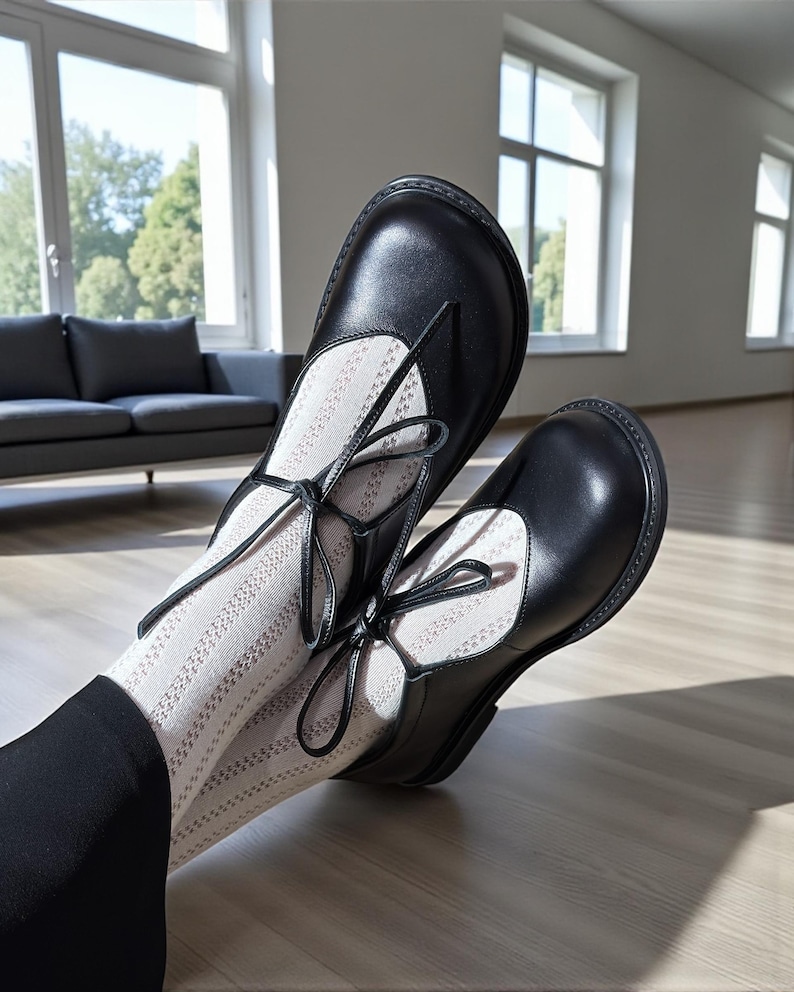 Op de afbeelding: Zwarte leren Mary Jane-schoenen met striksluitingen. De schoenen worden gedragen met witte sokken met een patroon. De foto is binnenshuis genomen met natuurlijk licht en een houten vloer.