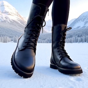 Könnte beinhalten: Schwarze Leder-Schnürstiefel mit seitlichem Reißverschluss und dicker Sohle. Die Stiefel werden mit schwarzen Leggings getragen. Der Hintergrund zeigt eine schneebedeckte Landschaft mit Bergen und blauem Himmel.