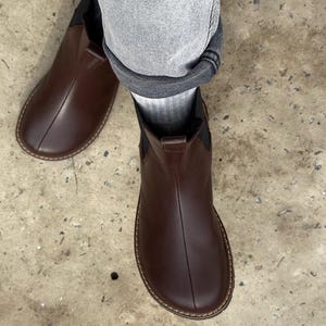May include: A pair of brown leather Chelsea boots. The boots have a rounded toe, elastic side panels, and a stitched sole. The boots are worn with gray denim jeans and gray socks. The boots are a dark brown color.