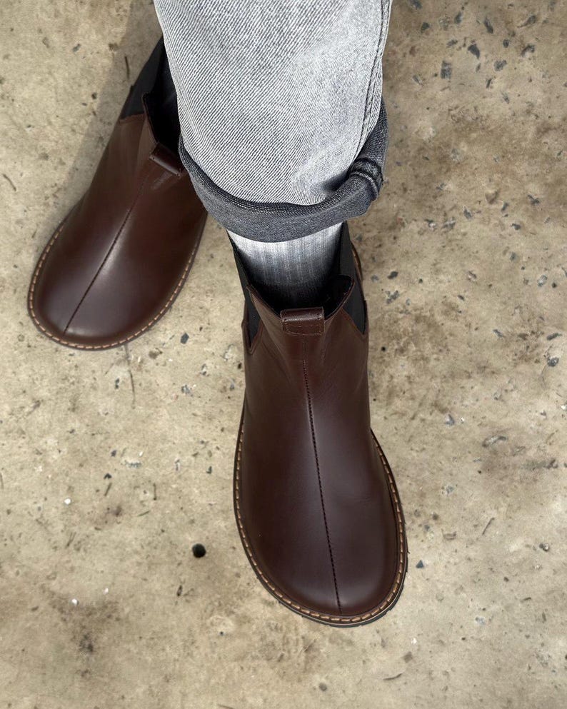 May include: A pair of brown leather Chelsea boots. The boots have a rounded toe, elastic side panels, and a stitched sole. The boots are worn with gray denim jeans and gray socks. The boots are a dark brown color.
