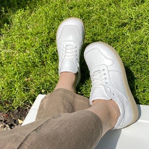 May include: A pair of white leather sneakers with white laces and a light brown sole. The shoes are worn by someone sitting on a white surface, with green grass in the background.