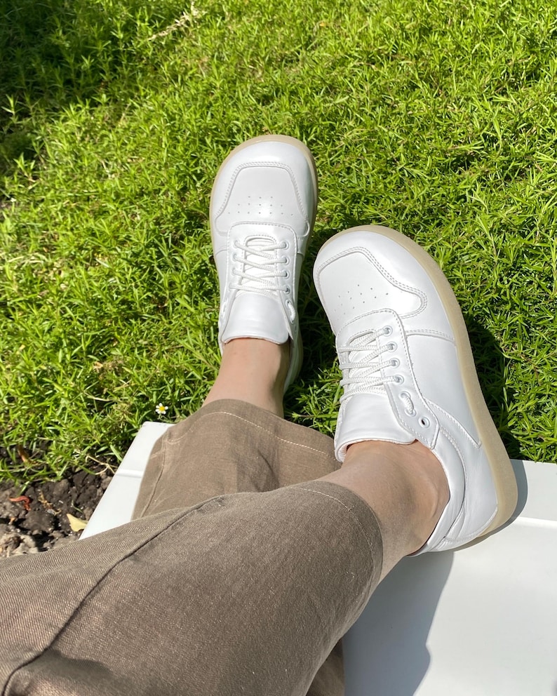 May include: A pair of white leather sneakers with white laces and a light brown sole. The shoes are worn by someone sitting on a white surface, with green grass in the background.