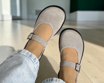 May include: A pair of light gray suede Mary Jane shoes featuring a rounded toe, a single strap across the instep with a small dark metal buckle, and a dark sole. The shoes are worn with light blue denim jeans, showing the top of the foot.