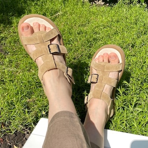 May include: Tan suede sandals with multiple straps and adjustable buckles. The sandals have a thick, light-colored sole and are worn outdoors on a sunny day. The sandals are a neutral color and appear to be comfortable.