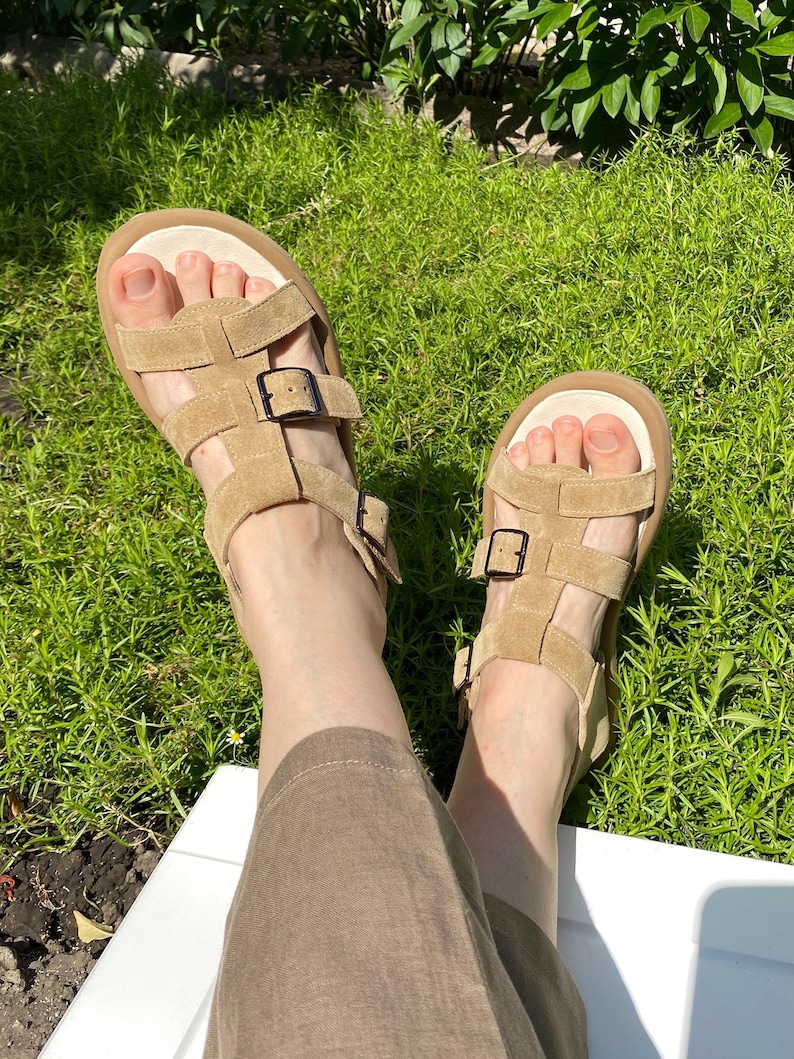 May include: Tan suede sandals with multiple straps and adjustable buckles. The sandals have a thick, light-colored sole and are worn outdoors on a sunny day. The sandals are a neutral color and appear to be comfortable.