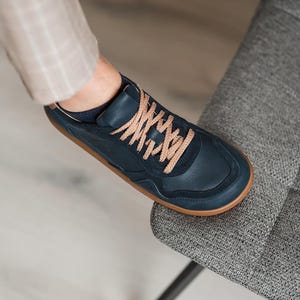 May include: Navy blue sneakers with tan laces and a brown sole. The shoe is made of leather and has a classic, minimalist design. The shoe is on a gray chair.