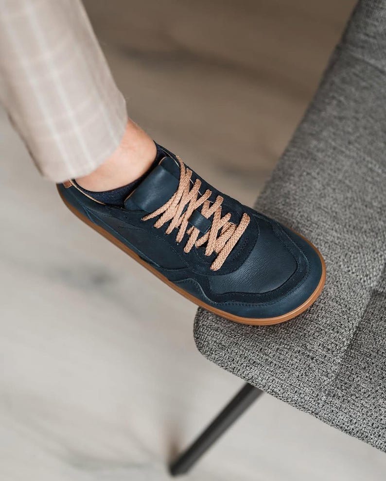 May include: Navy blue sneakers with tan laces and a brown sole. The shoe is made of leather and has a classic, minimalist design. The shoe is on a gray chair.