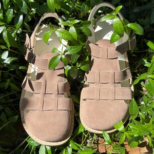 May include: A pair of tan suede sandals with a woven design on the upper. The sandals have adjustable ankle straps with buckles and are set against a backdrop of green foliage.