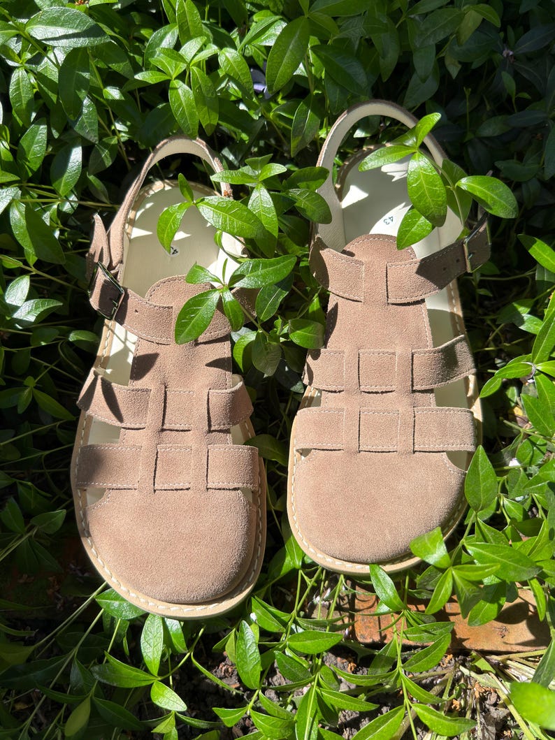 May include: A pair of tan suede sandals with a woven design on the upper. The sandals have adjustable ankle straps with buckles and are set against a backdrop of green foliage.