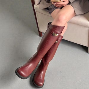 May include: A pair of knee-high boots in a deep burgundy color, featuring a smooth finish, a rounded toe, and a decorative gold-toned metal buckle strap detail near the top of the shaft. The boots have a thick, dark-colored rubber sole.