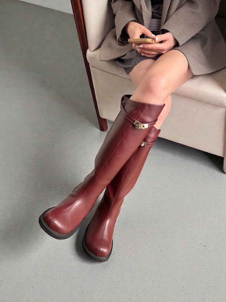 May include: A pair of knee-high boots in a deep burgundy color, featuring a smooth finish, a rounded toe, and a decorative gold-toned metal buckle strap detail near the top of the shaft. The boots have a thick, dark-colored rubber sole.