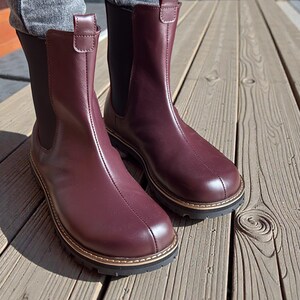 May include: A pair of burgundy leather Chelsea boots. The boots have a rounded toe, a stitched seam down the front, and a gold-colored trim around the sole. The boots have a black elastic side panel and a pull tab at the top.