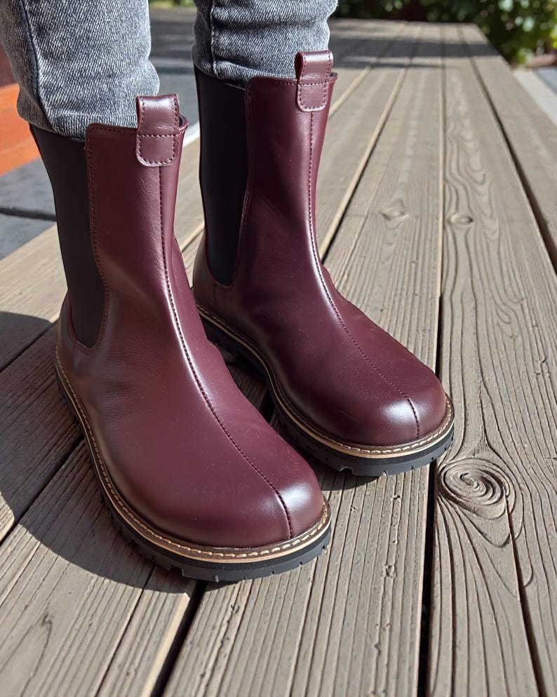 May include: A pair of burgundy leather Chelsea boots. The boots have a rounded toe, a stitched seam down the front, and a gold-colored trim around the sole. The boots have a black elastic side panel and a pull tab at the top.