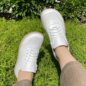 May include: White leather sneakers with a low-top design. The shoes have white laces and a light-colored sole. The sneakers are shown on a background of green grass and foliage.