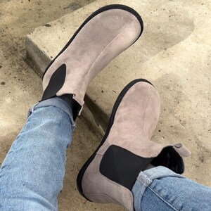 May include: A pair of light gray suede ankle boots with black elastic side panels and black soles. The boots are worn with blue jeans.