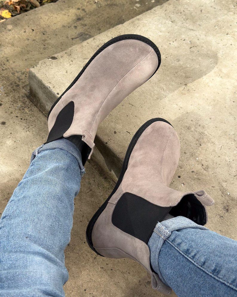 May include: A pair of light gray suede ankle boots with black elastic side panels and black soles. The boots are worn with blue jeans.