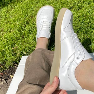 May include: A pair of white sneakers with a low profile and a light brown sole. The sneakers have a classic lace-up design and are made of a smooth, shiny material. The shoes are shown on a person's feet.