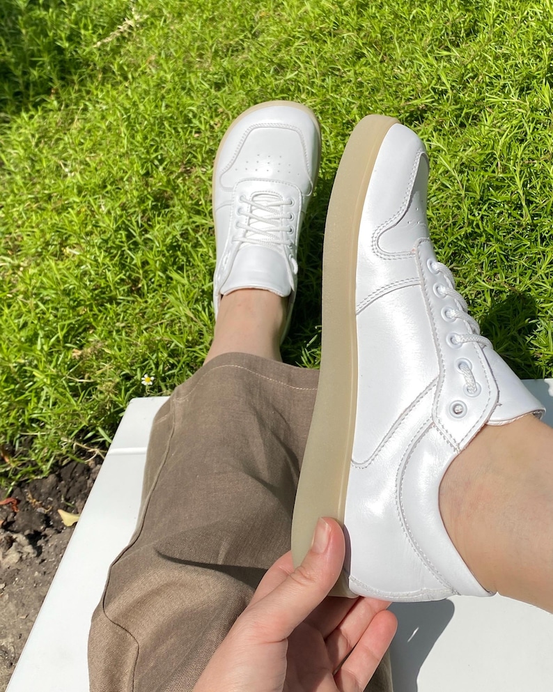 May include: A pair of white sneakers with a low profile and a light brown sole. The sneakers have a classic lace-up design and are made of a smooth, shiny material. The shoes are shown on a person's feet.