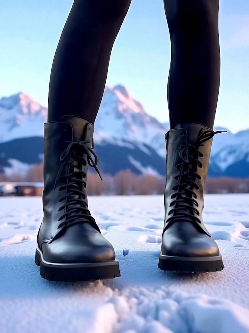 May include: Black lace-up boots with a lug sole. The boots are worn with black leggings, standing in the snow. The background features snow-covered mountains and a blue sky.