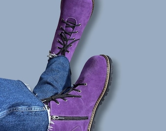 May include: A pair of vibrant purple suede boots featuring black laces, silver-toned eyelets, and a side zip closure. The boots have a rounded toe, a thick black rubber sole with visible stitching, and are worn with blue denim jeans.