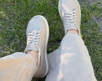 May include: A pair of beige sneakers with white soles and laces. The shoes are worn with beige pants, and the image is taken from a first-person perspective, with green grass in the background.