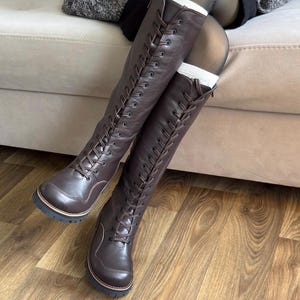 May include: Dark brown knee-high boots featuring a full-length front lace-up design with matching brown laces, a rounded toe cap, and a thick black lug sole. The boots extend to the knee and include a side zipper closure for easy access.