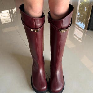 May include: A pair of knee-high boots in a deep burgundy color, crafted from a smooth material. Each boot features a thin strap encircling the upper shaft, secured with a small, rectangular gold-toned metal buckle detail at the front.