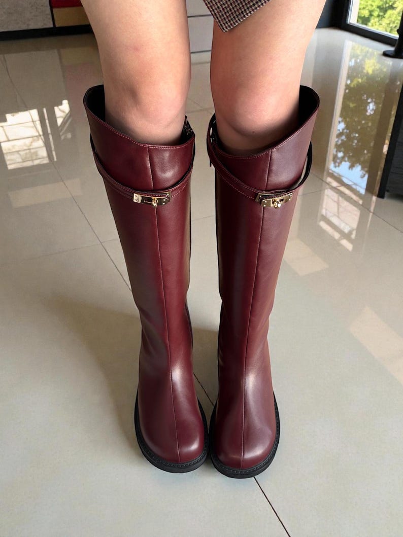 May include: A pair of knee-high boots in a deep burgundy color, crafted from a smooth material. Each boot features a thin strap encircling the upper shaft, secured with a small, rectangular gold-toned metal buckle detail at the front.