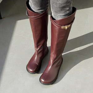 May include: A pair of knee-high boots in a deep burgundy color, crafted with a smooth finish. Each boot features a thin strap encircling the upper shaft, secured with a polished gold-tone metal buckle hardware detail on the outer side.