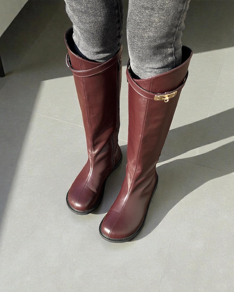 May include: A pair of knee-high boots in a deep burgundy color, crafted with a smooth finish. Each boot features a thin strap encircling the upper shaft, secured with a polished gold-tone metal buckle hardware detail on the outer side.
