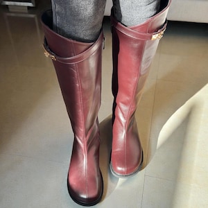 May include: A pair of knee-high boots in a deep burgundy leather finish. The boots feature a smooth, rounded toe design and a decorative strap across the upper shaft secured with a gold-toned metal buckle detail on the outer side of each boot.