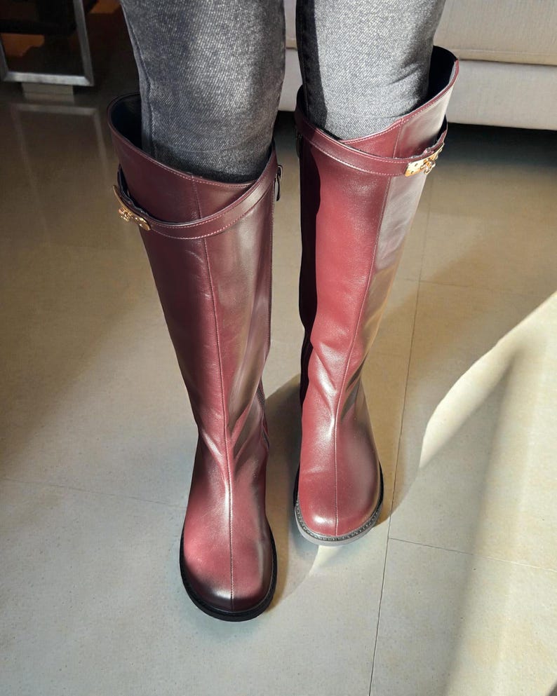 May include: A pair of knee-high boots in a deep burgundy leather finish. The boots feature a smooth, rounded toe design and a decorative strap across the upper shaft secured with a gold-toned metal buckle detail on the outer side of each boot.