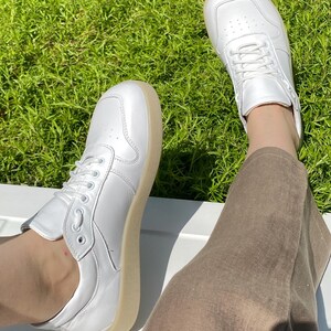 May include: A pair of white sneakers with a light brown sole. The sneakers have a classic design with laces and are made of a smooth material. The shoes are shown on a white surface with green grass in the background.