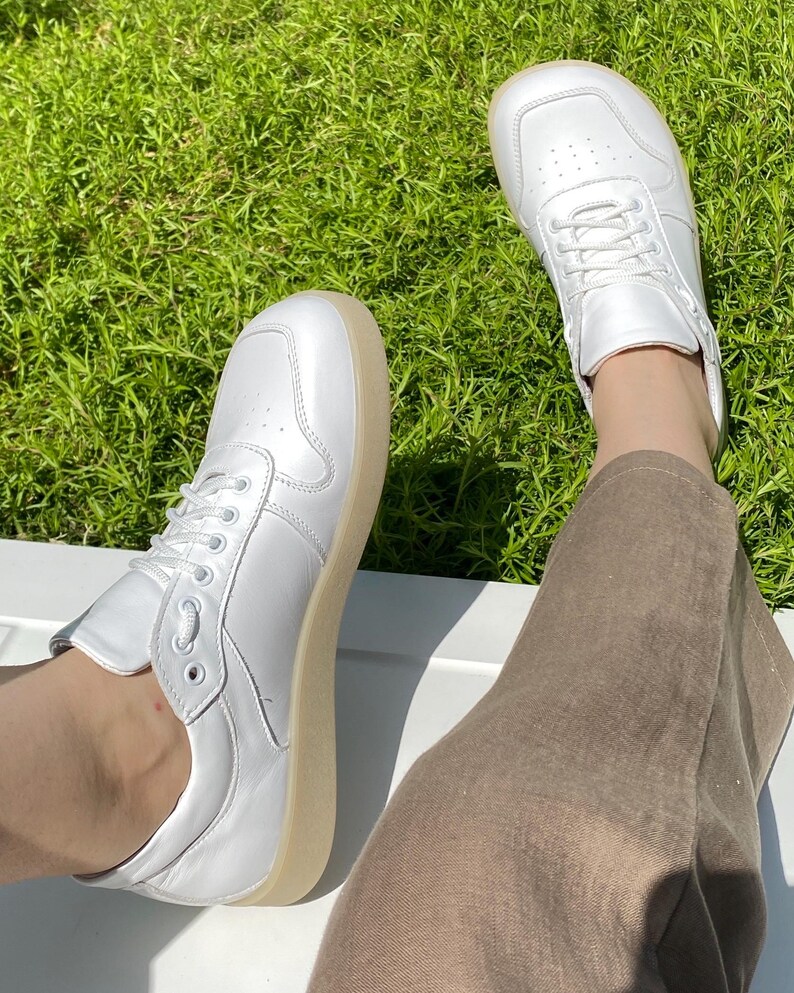 May include: A pair of white sneakers with a light brown sole. The sneakers have a classic design with laces and are made of a smooth material. The shoes are shown on a white surface with green grass in the background.