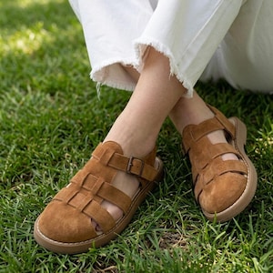 May include: Brown suede sandals with a woven design and a buckle closure. The sandals have a round toe and a thick, light brown sole. The shoes are shown on a background of green grass.