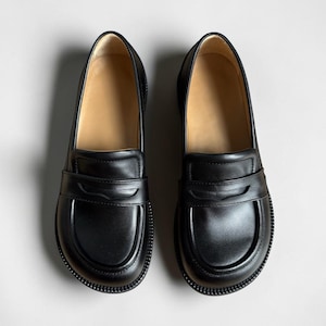 Puede incluir: Un par de mocasines de cuero negro con punta redondeada, una tira cl&aacute;sica tipo penny sobre el empeine y una suela gruesa de goma texturizada. El interior de los zapatos est&aacute; forrado con un material de color tostado claro y el exterior es liso.