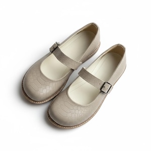 May include: A pair of taupe Mary Jane style shoes with a textured, crocodile-like pattern. Each shoe has a strap with a small buckle closure. The shoes have a rounded toe and a light-colored interior.