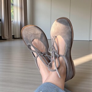 Op de afbeelding: Een paar grijze su&egrave;de ballerina's met een ronde neus en een vetersluiting. De schoenen hebben een lichtbruine zool. De veters zijn in een strik geknoopt. Gepresenteerd op een lichte houten vloer.