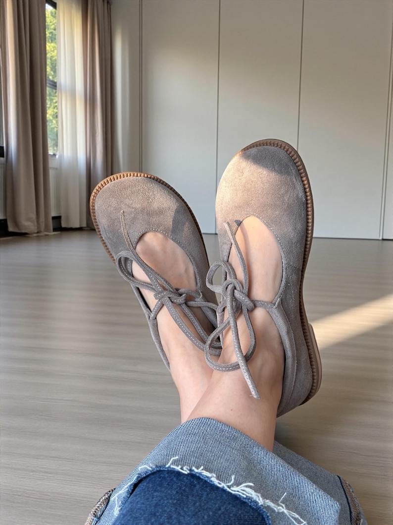 Op de afbeelding: Een paar grijze su&egrave;de ballerina's met een ronde neus en een vetersluiting. De schoenen hebben een lichtbruine zool. De veters zijn in een strik geknoopt. Gepresenteerd op een lichte houten vloer.