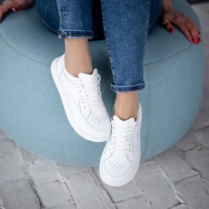 May include: White trainers with a low-profile design. The shoes have a lace-up closure and a perforated pattern on the toe box. The trainers are paired with blue jeans. The person is sitting on a light blue ottoman.