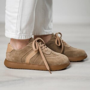 May include: A pair of light brown suede sneakers with white laces and a brown rubber sole. The sneakers have a light brown stripe across the side.