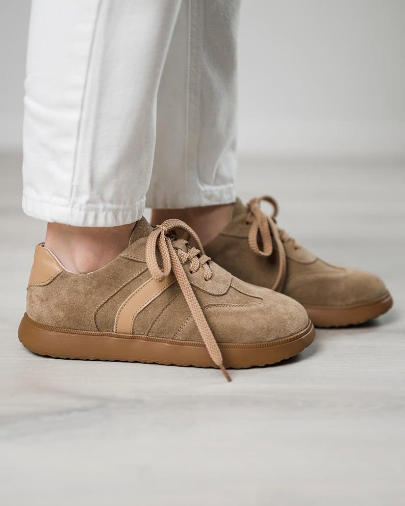May include: A pair of light brown suede sneakers with white laces and a brown rubber sole. The sneakers have a light brown stripe across the side.