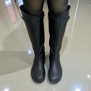 May include: A pair of black knee-high boots featuring a smooth matte finish, a rounded toe, a vertical seam running down the center of the shaft, and a thin horizontal strap detail with a small buckle positioned near the top of the boot shaft.